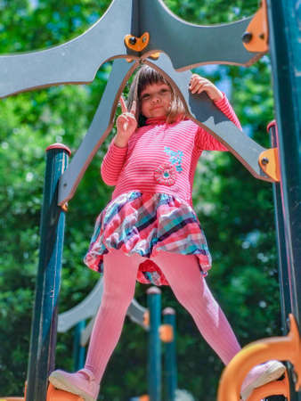 Girl in a pink dress playing on top of a playground while looking at the camera and making the...
