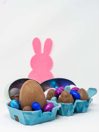 Lots of chocolate Easter eggs of different sizes in an egg carton and in egg-shaped containers on a white background with a silhouette of a pink bunny.の写真素材