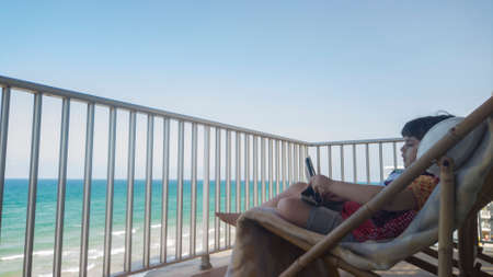 Little girl sitting on a deck chair on a terrace with sea view playing with a tabletの写真素材