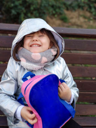 Back-to-school elementary girl in silver hooded coat and scarf smiling on a bench while holding her...