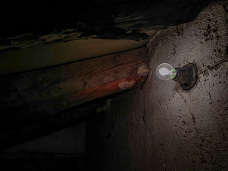 Light bulb glowing dimly in an abandoned old basement with wooden beams and stone wallの写真素材