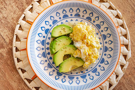 A plate of scrambled eggs served alongside slices of fresh avocado on top of a piece of corn flour...