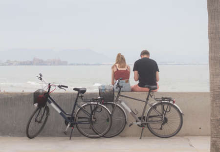 A couple of cyclist tourists rest seated observing the views of the beach