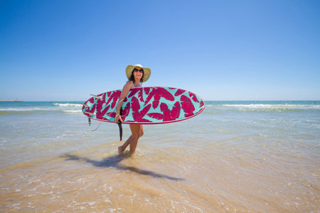 Attractive happy middle-aged woman wearing a hat, sunglasses and carrying a surfboard on the beachの写真素材