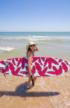 Attractive happy middle-aged woman wearing a hat, sunglasses and carrying a surfboard on the beachの写真素材