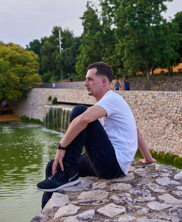 A young, attractive boy sitting on a stone wall next to a pond.の写真素材
