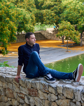 A young, attractive boy sitting on a stone wall next to a pond.の写真素材