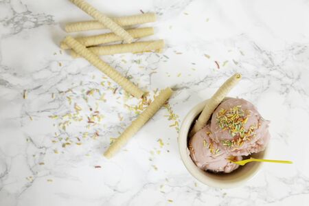 Two strawberry ice cream balls on ceramic bowl with yellow plastic teaspoon and biscuit and various biscuits on marble background from above. Copy space. Top viewの写真素材