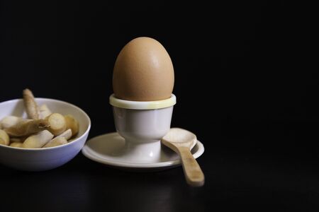 soft boiled egg in eggcup, wooden spoon and ceramic bowl with breadsticks on black background, Copy spaceの写真素材