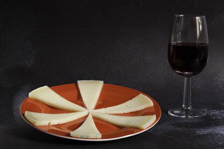 six slices of typical spanish manchego cheese on red ceramic plate and red wine glass on dark background. Copy spaceの写真素材