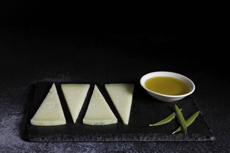 four slices of typical spanish manchego cheese and bowl olive oil on slate plate and dark background. Copy spaceの写真素材
