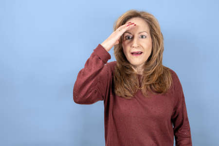 Woman with a surprised face and hand on the forehead for emotion, blue studio background.の写真素材