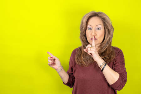 Woman pointing at the wall and with her finger on her mouth asking for silence, yellow studio...