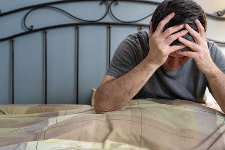 Man with insomnia sitting on the bed with his hands on his head and being exhausted.