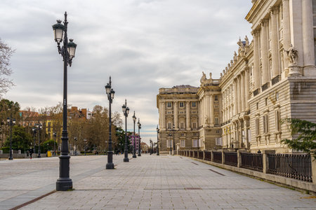 Side facade of the royal palace of Madrid, pedestrian street with lampposts, trees and sunny day with clouds. Spain.のeditorial素材