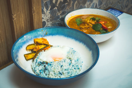 a bowl of curry chicken with potatoes and butterfly pea rice and pickled achar and fried eggの写真素材