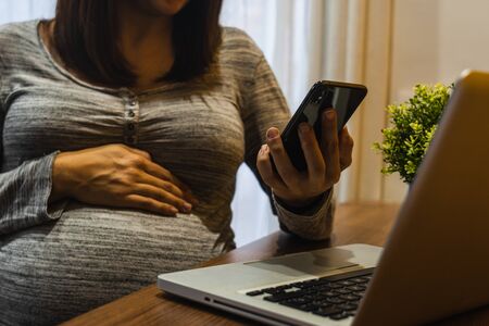 Pregnant white woman working with a laptop and phone on a wood tableの写真素材
