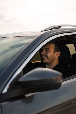Man sitting on the seat of a new car, smiling while looking forward, wearing dark clothes. Caucasian man with dark hair and a short beard. In the evening in a dark gray car.の写真素材