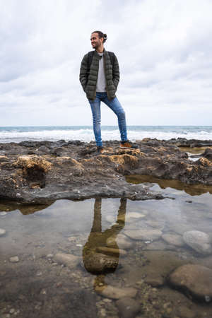 Happy man reflected in a puddle in the sea. On a cloudy day. He is wearing a green jacket, gray sweater and jeans. Caucasian, with ponytail and rucksack on his back.の写真素材