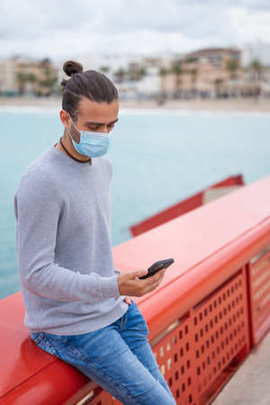 Man using mobile phone with surgical mask leaning on a red railing. He is on a promenade, on a cloudy day. He has one hand in his pocket. He is wearing jeans, a gray jumper. Alicante, Javeaの写真素材