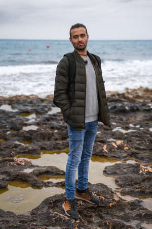 Man posing in the sea with attitude and a backpack. In a sea of rocks surrounded by puddles on a cloudy day. He is wearing a backpack, jeans, sweater and green jacket. His hands are in his pockets.の写真素材