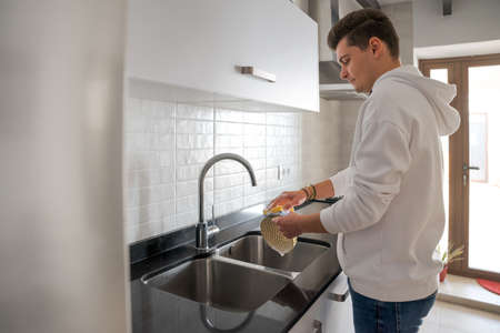 Caucasian brown-haired man in the kitchen. He is soaping the dishes. In a white kitchen with black...