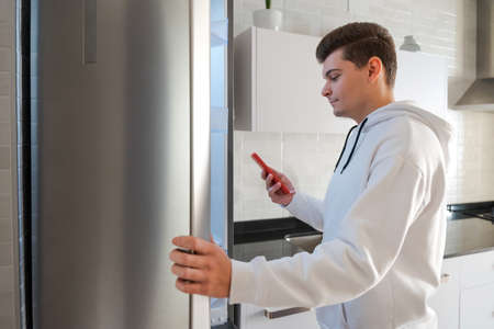 Caucasian man in the kitchen making the shopping list. He is in front of the fridge checking the list on his mobile phone. He is dressed in a white sweatshirt and his mobile phone is red. Spainの写真素材