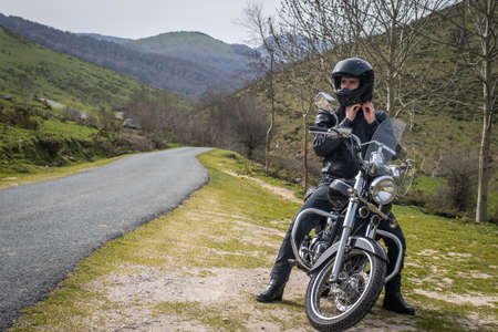 Young biker putting on his black helmet while sitting on his custom motorcycle next to a mountain roadの写真素材