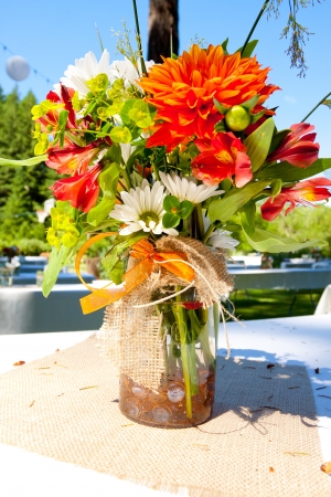 White, orange, and green wedding flower arrangements with burlap as the centerpieces for table settings at an outdoor reception.の写真素材