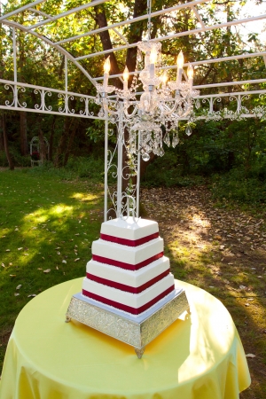 A white and red square wedding cake with multiple tiers and the letter R on top sits outside on a yellow table for a wedding reception の写真素材