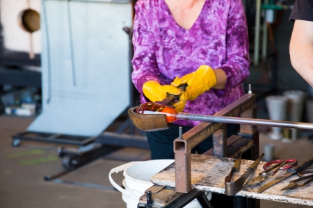 A woman is shaping some motlen glass into a bowl using a wooden forming tool at a glass studio.の写真素材