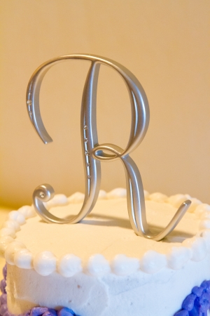A cake topper with the letter R on it on this white and purple cake indoors. A cake topper with the letter R on it on this white and purple cake indoors.