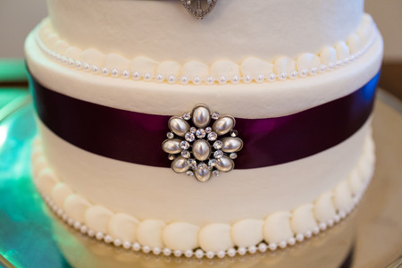 Tall white wedding cake wrapped in purple ribbon with brooches attached to each tier of the dessert.の写真素材