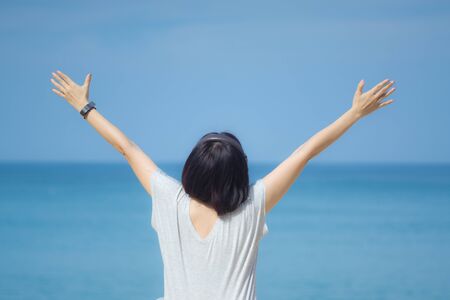 Young asian woman in a gray dress holding hands up to the sky. Blue sky and crystal sea of tropical beach as background. Photo from behind. Enjoy the summer time. Happy island lifestyle. Concept for relaxation, travel, vacation and holidays with copy space.の写真素材