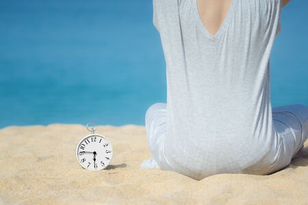 Asian women in a gray dress sitting beside the white alarm clock placed on the sand. Blue sea and sky as a background. Concept for relaxation, travel vacation and holidays with copy space. Summer time.の写真素材