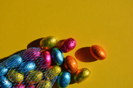 Foil wrapped chocolate Easter eggs spilling out of blue plastic net bag, isolated on bright yellow background with copy spaceの写真素材