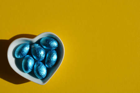White heart shaped dish with blue foil wrapped chocolate Easter eggs isolated on bright yellow background with copy spaceの写真素材