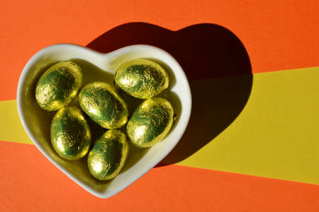Gold foil wrapped chocolate Easter eggs in a heart shaped dish isolated on yellow and orange backgroundの写真素材