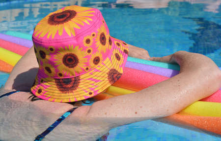Woman wearing sun hat floating on swim noodles in swimming pool, from behindの写真素材