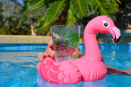 Refreshing drink in woman's hand, in bright pink flamingo glass and holder floating in swimming poolの写真素材