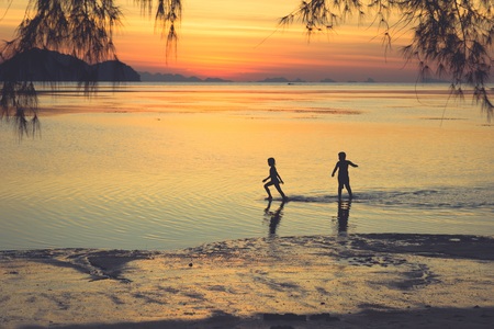 Little girl walking away from her boyfriend on shallow water at sunset on the beach and tree...