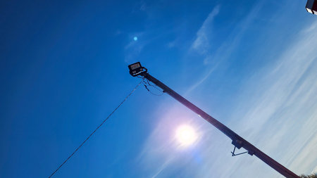 A tall wooden utility pole with a floodlight rises against a clear blue sky, sunflare visible near the pole. Ground-level view reveals trees and a small building in a rural setting.の写真素材