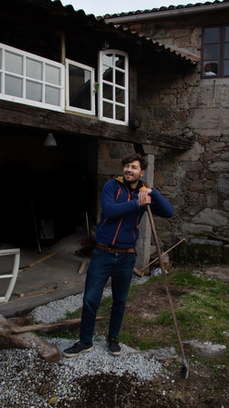 Smiling young man with blue clothes leaning on a rake in a country house garden