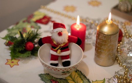 Cute Santa Claus doll inside a bowl with on a Christmas tablecloth with candlesの写真素材