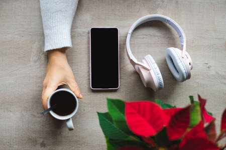 Top view of a table with a poinsettia, wireless headphones, mobile phone and a hand grabbing a cup of coffeeの写真素材
