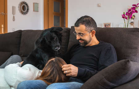 Young happy couple on the sofa at home with their black dog. Woman resting her head on the legs of her partner.の写真素材