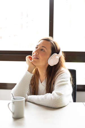 Happy and relaxed woman uses her wireless headphones at home leaning on the table where there is a cupの写真素材