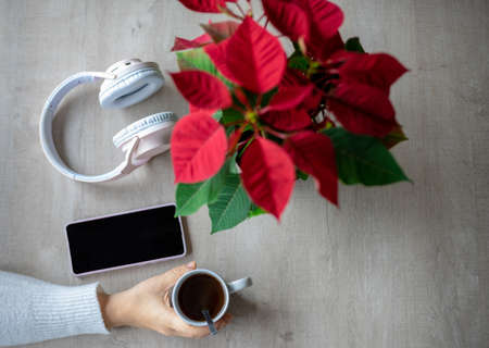 Top view of a table with a poinsettia, wireless headphones, mobile phone and a hand grabbing a cup of coffeeの写真素材