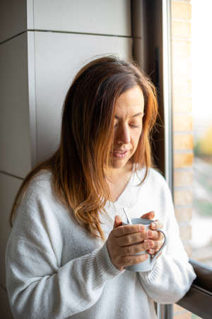 Young woman watches from her home window while holding a hot mugの写真素材