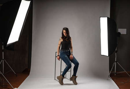 Alternative model with tattoos and jewelry poses sitting in a chair in a professional photo studio with spotlights and white backgroundの写真素材